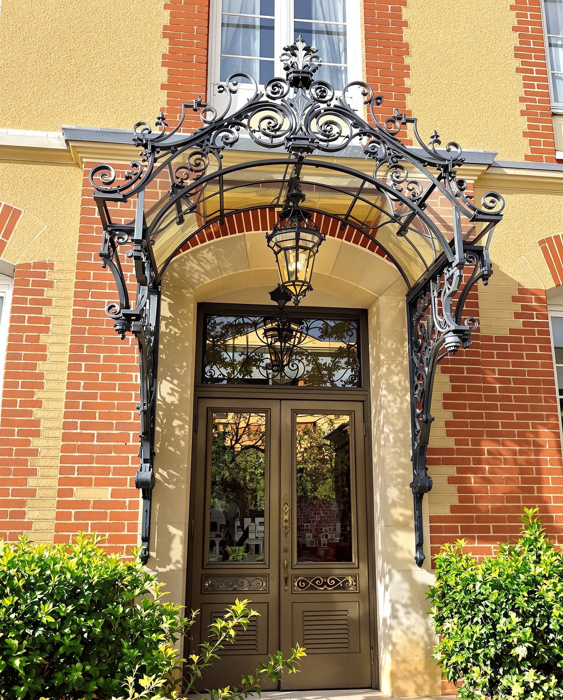 Façade d'une élégante maison meulière typique de l'Île-de-France, avec ses pierres aux teintes chaudes de jaune et d'orangé, des détails en brique rouge et une marquise en fer forgé ouvragé au-dessus de la porte d'entrée.